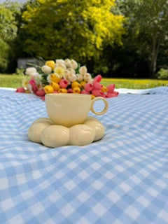 Tasse jaune pastel design en forme de nuage, lors d'un pique-nique en extérieur sur une nappe vichy bleue, avec jardin verdoyant en arrière-plan 