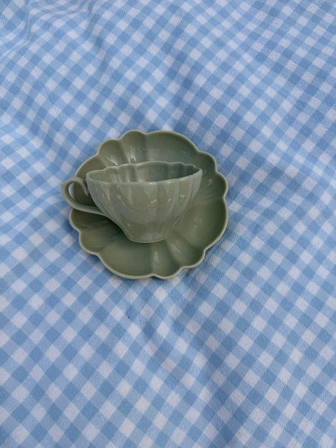 Tasse en céramique vert kaki avec soucoupe en forme de fleur, photographiée de dessus sur une nappe à carreaux vichy bleu clair.