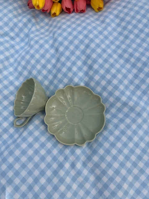 Tasse en céramique vert kaki avec soucoupe en forme de fleur, photographiée de dessus sur une nappe à carreaux vichy bleu clair.