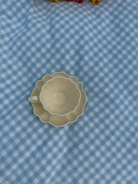 Tasse en céramique crème avec soucoupe en forme de fleur, photographiée de dessus sur une nappe à carreaux vichy bleu clair.