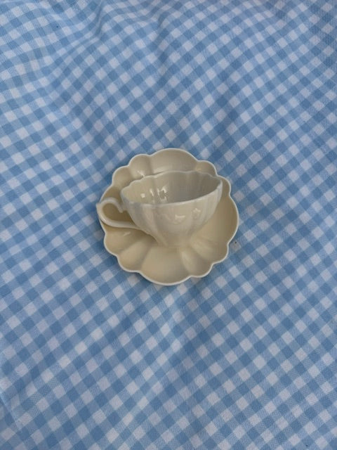 Tasse en céramique crème avec soucoupe en forme de fleur, photographiée de dessus sur une nappe à carreaux vichy bleu clair.