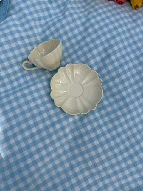 Tasse en céramique crème avec soucoupe en forme de fleur, photographiée de dessus sur une nappe à carreaux vichy bleu clair.